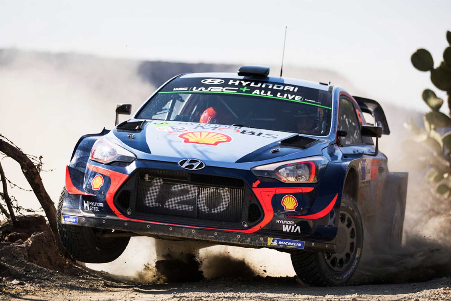 Hyundai i20 WRC impulsado por Shell compitiendo en una etapa de rally sobre terreno arido levantando polvo entre cactus y rocas