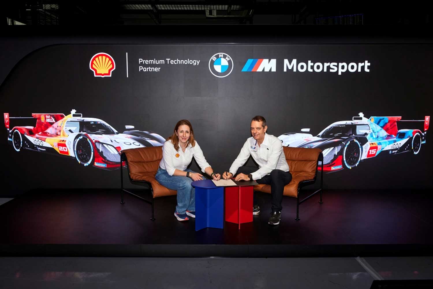 Representantes de Shell y BMW Motorsport sellando su alianza tecnologica frente a un fondo con prototipos de competicion y logotipos oficiales